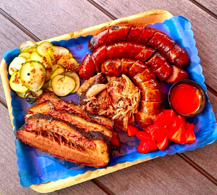 A paper-lined tray showcases thick slices of brisket, three types of sausage, pickles, strawberries, and sauce, all set on a rustic wooden table.