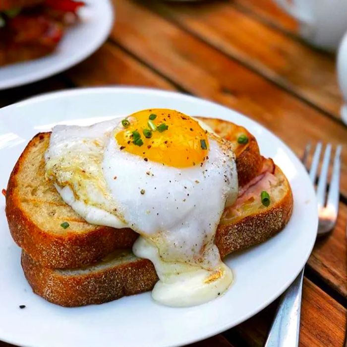 A croque madame with a perfectly runny egg, served on a plate next to a fork on a rustic wooden patio table.