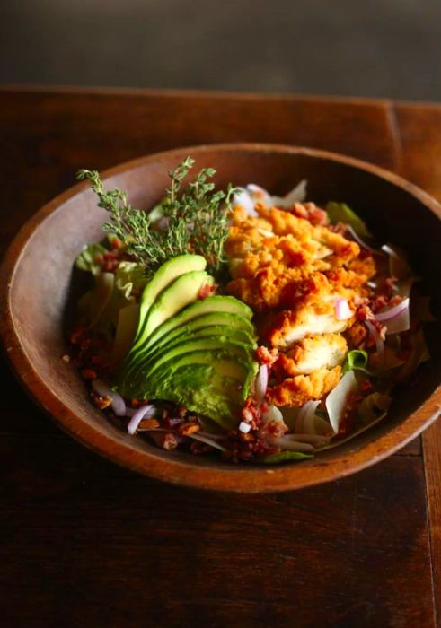 A deep wooden bowl holds a vibrant array of sliced fried chicken, ripe avocado, fresh rosemary sprigs, and assorted vegetables, all set on a rustic wooden table.