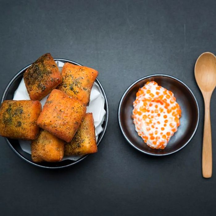 An aerial view of puffed square donuts sprinkled with herb salt, accompanied by a bowl of creamy sauce flecked with roe, set on a dark surface with a wooden spoon.