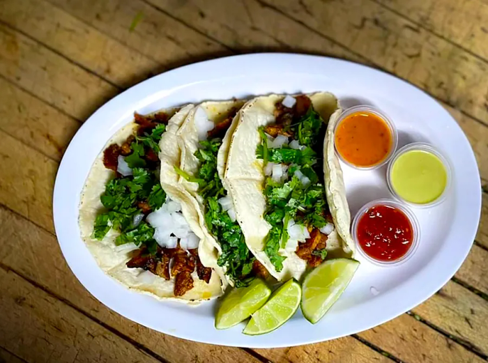 A plate of three tacos filled with meat, diced onions, and cilantro, accompanied by lime wedges and an array of sauces.