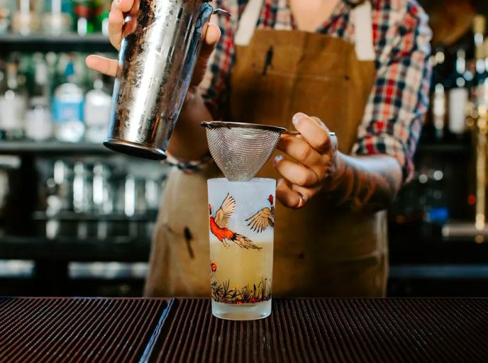 A bartender pours a cocktail into a highball glass adorned with illustrations of flying birds, set against a blurred backdrop of bottles and glasses.