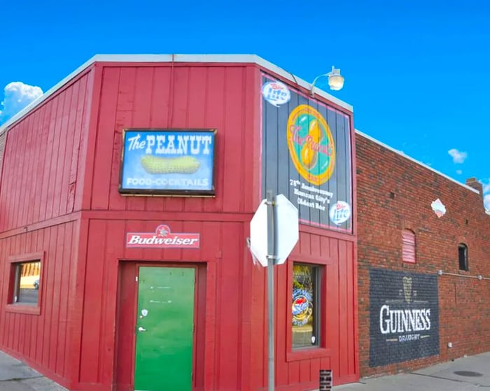 The exterior of a bar featuring red wood and brick, with signage for The Peanut showcasing illustrations of peanuts in their shells, alongside advertisements for various alcohol brands.