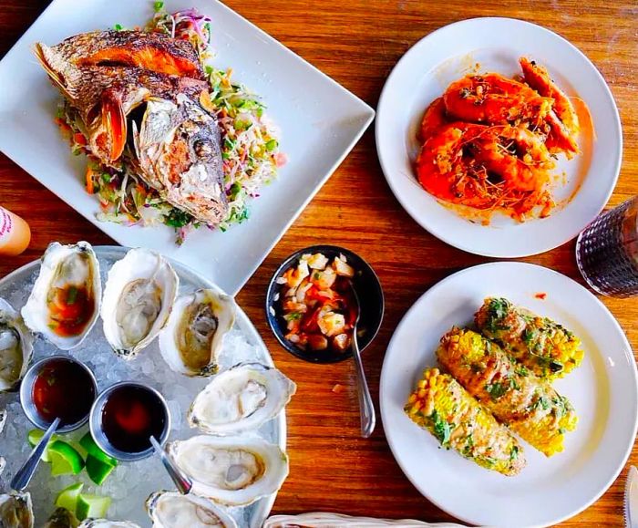 A table overflowing with fresh oysters on ice, sauced and herb-topped corn, succulent grilled shrimp, and a beautifully prepared fish head.