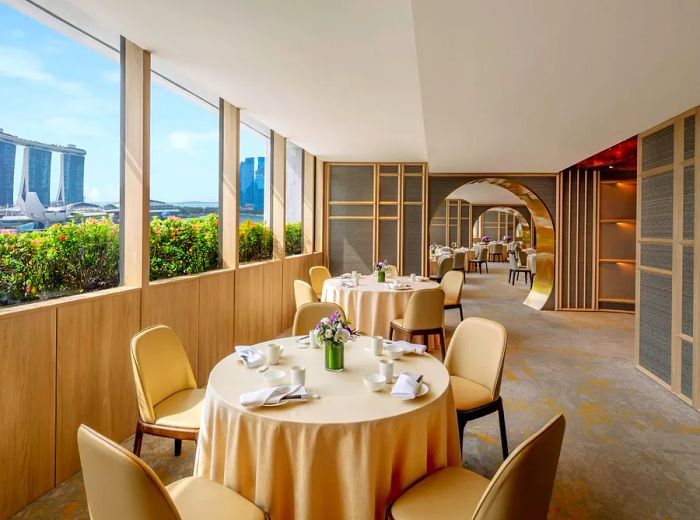 A luminous dining room perched high above, offering a panoramic view of a tree-lined street. The tables are adorned with cream-colored tablecloths, while the gray-paneled walls create a sophisticated backdrop. Further seating areas are visible through elegantly rounded archways.