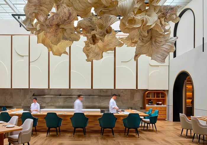 An expansive, stylish restaurant with chefs working behind a central counter, beneath a large, elaborate mobile installation.