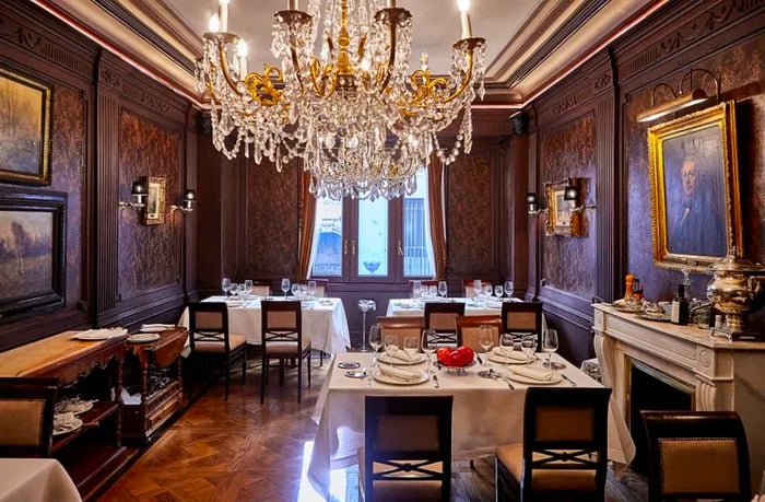 The dining room exudes sophistication with its dark wooden walls, a sparkling crystal chandelier, crisp white tablecloths, and elegant classical artwork.