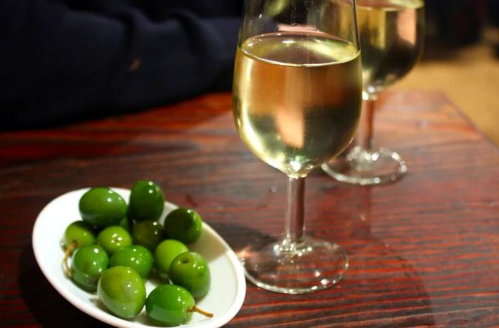 A glass of sherry sits beside a small plate of olives on a wooden bar, with someone leaning casually nearby.