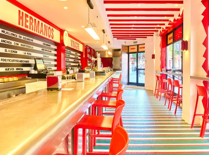 The interior of the restaurant is a bright mix of white and red, featuring vibrant red stools, a beamed ceiling, a long counter, and expansive windows.