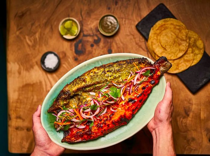 Viewed from above, a plate holds a whole cooked fish with green marinade on one side and red on the other, garnished with sliced vegetables. Surrounding the plate on a wooden table are tortillas, lime wedges, salt, and extra sauce.