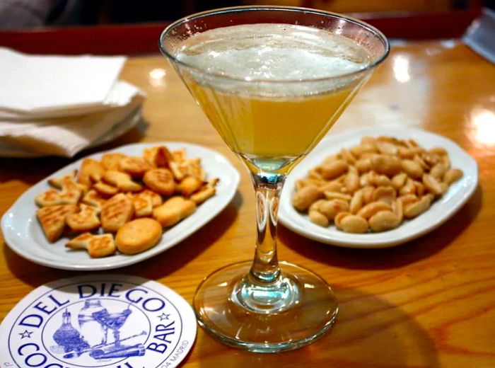 A martini glass filled with a cocktail is surrounded by small plates of crunchy snacks and a coaster marked with the Del Diego Cocktail Bar logo.