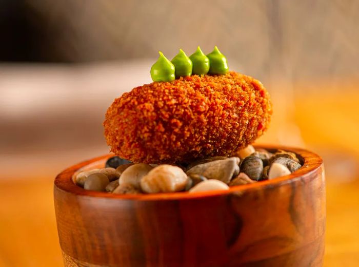 A close-up of a croquette resting on a bed of stones inside a wooden bowl, garnished with vibrant green sauce.