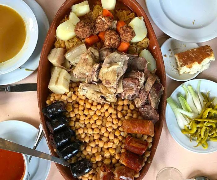 A large platter displays an assortment of meats and chickpeas surrounded by various half-eaten side dishes, including vegetables, breads, and soups.