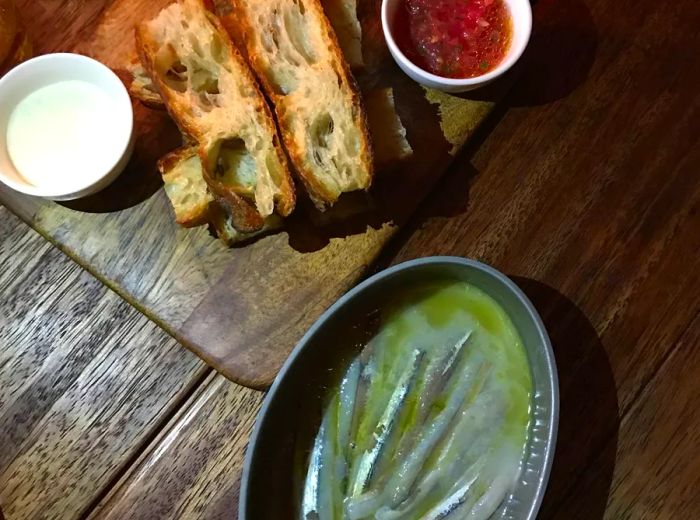 A bowl of smelt in sauce served alongside a bread board with avocado slices and tomato sauce.