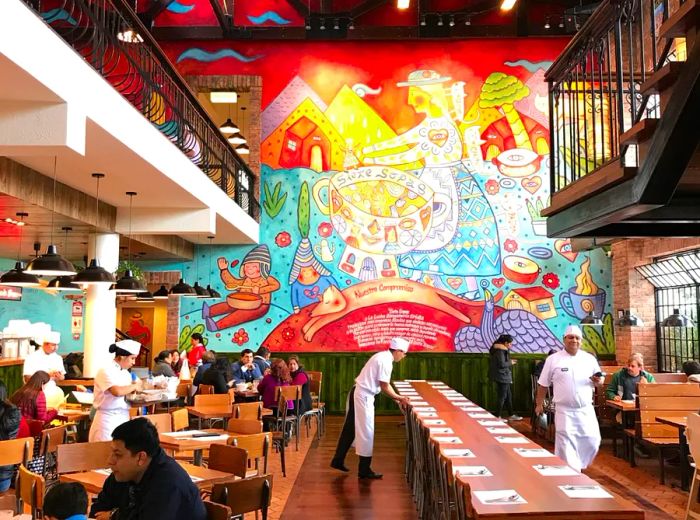 The two-story dining area features a spiral staircase partially out of sight. A vibrant mural of a woman making soup adorns the tall back wall. Families gather around four-top tables while a long communal table is being set up in the center.