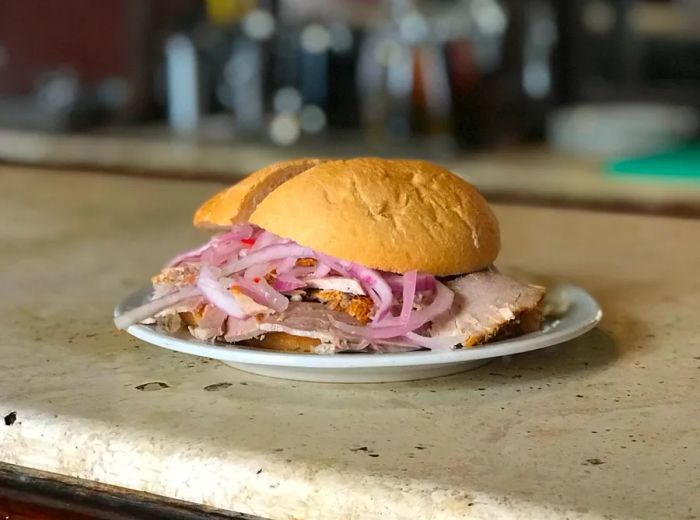 A plate holds a generously stuffed sandwich with pork and red onions, left alone on a bar counter.