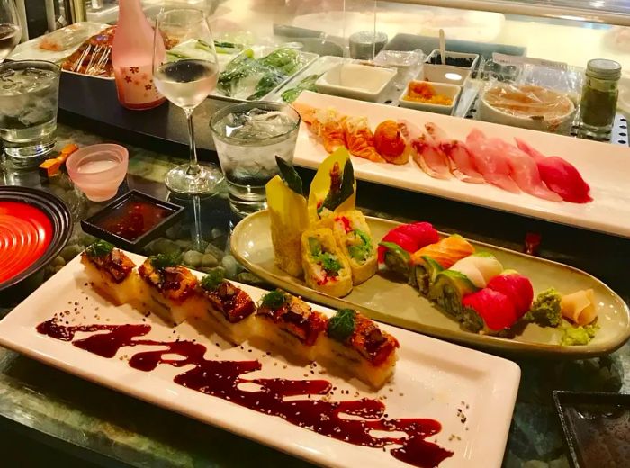 An array of beautifully crafted maki rolls and nigiri, paired with sake and wine, displayed before a sushi counter where the chef prepares dishes.