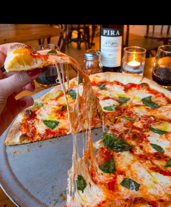 A slice of pizza is being lifted from a whole pie on a platter, with gooey cheese stretching, set next to a bottle of wine and a pepper shaker.