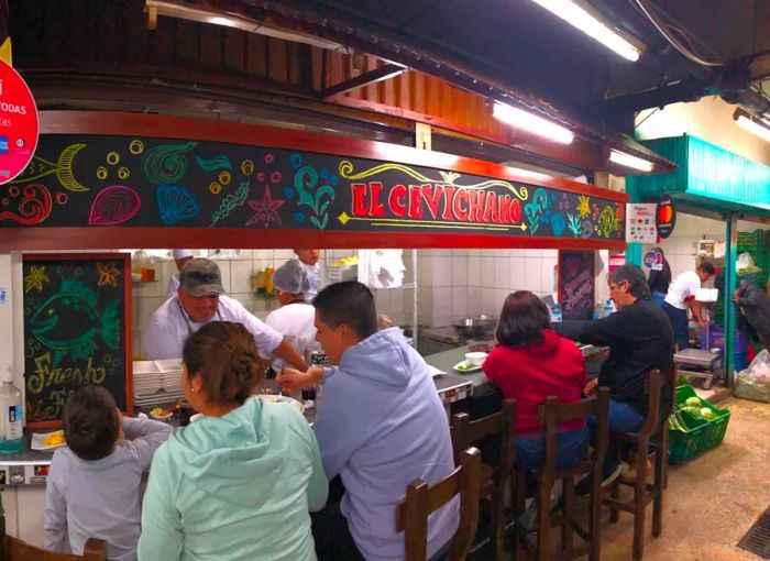 Diners enjoy their meals at a counter under a vibrant sign for El Cevichano, as chefs pass plates of food from the kitchen.