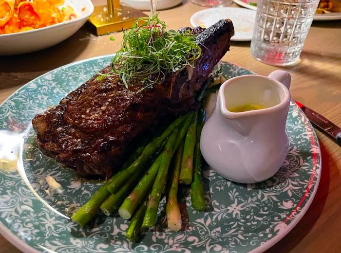 A hearty bone-in steak served alongside asparagus and a dish of rich sauce.