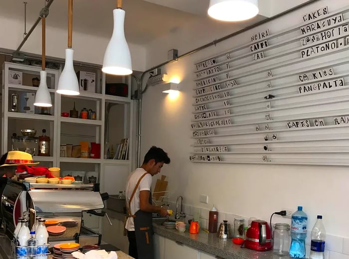 A marble counter showcases jars of preserves and coffee bags, with an espresso machine at the bar's far end. A barista prepares dishes behind the counter, and a letter board lists menu items on the back wall.