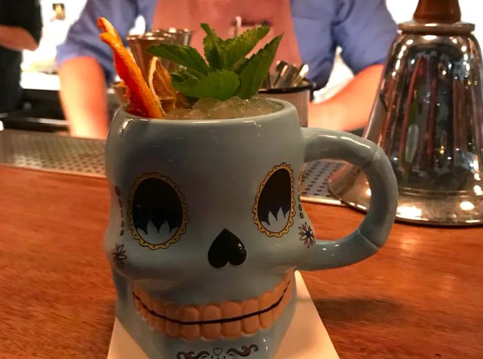 A cocktail served in a quirky skull mug, adorned with a cinnamon stick, fresh herbs, and topped with crushed ice that extends above the rim.