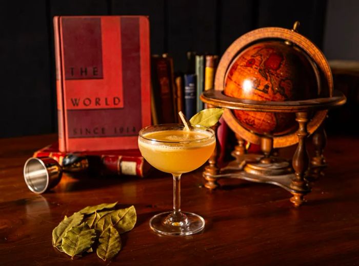 An orange cocktail in a coupe glass sits among a backdrop of a globe, historical book, cocktail jigger, and bay leaves.