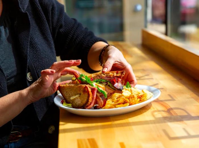 A diner dissects a large sandwich on a sunny windowsill, with a side of potato chips.