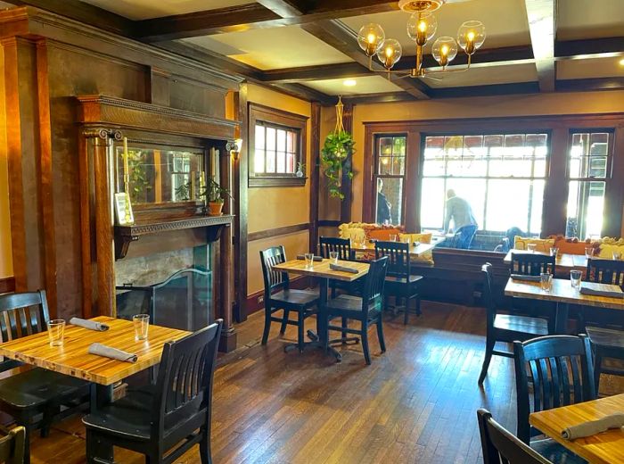 A dining area featuring a rustic wood-beam ceiling adorned with a chandelier, complemented by a grand fireplace, wooden flooring, and tables arranged for an elegant dinner setting.