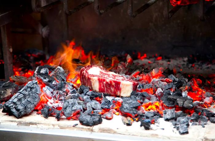 A substantial piece of marbled meat sizzles directly over a charcoal fire