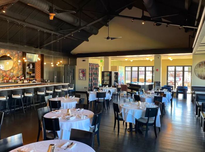 A spacious dining area with high ceilings, tables set with white tablecloths, a bar running along one wall, and tall windows at the end of the room.