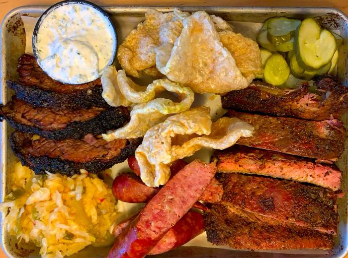 A metal tray lined with paper, loaded with brisket slices, ribs, sausages, cracklings, coleslaw, cucumbers, and dipping sauce.