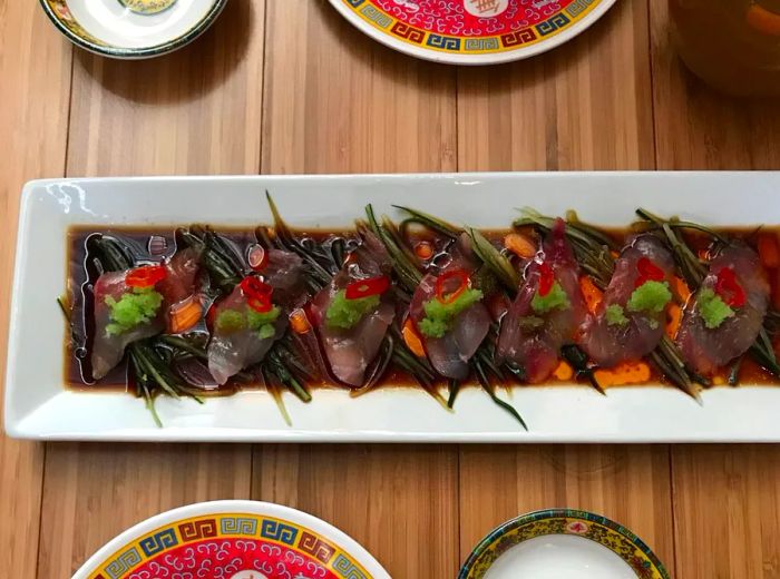 A platter featuring slices of cured fish drenched in ponzu and sesame oil, garnished with Japanese cucumber and limo chile slices.