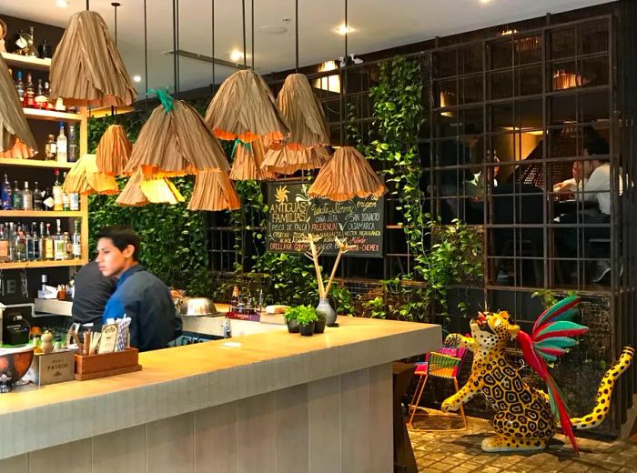 A bar counter adorned with a cluster of pendant lights at varying heights, bar shelves on one side, a jaguar statue and a vibrant jungle bird on the floor, and a lush vine-covered wall in the backdrop.