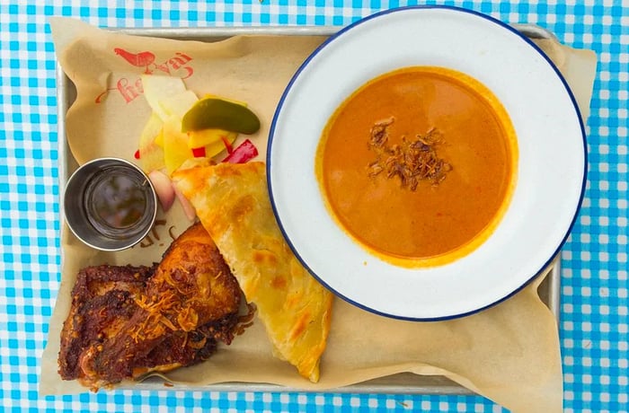 An aerial view of a metal tray featuring a bowl of orange curry, folded roti, and Thai-style fried chicken.