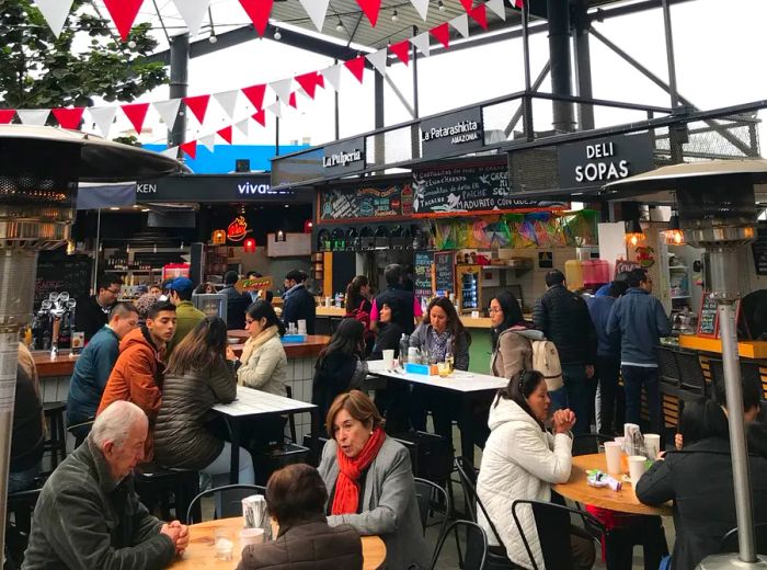 An expansive, warehouse-style venue flooded with natural light through the roof and high wall panels, adorned with flags suspended above diners and food stalls in the background.