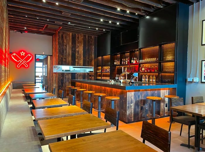 An interior shot of a barbecue restaurant featuring tables, a bar, and a neon sign with crossed cleavers.