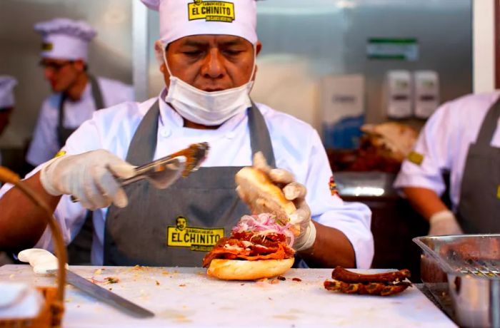 A server in a branded apron and chef's hat lifts the top bun of a sandwich while using tongs to add the filling.