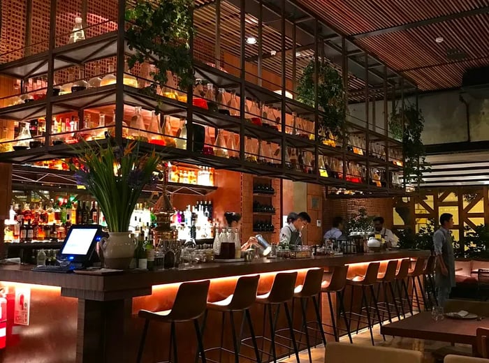 A centrally located bar at Mayta, featuring a dark ambiance with industrial steel shelves above and illuminated bar stools and bottles, creating a sleek and modern look.