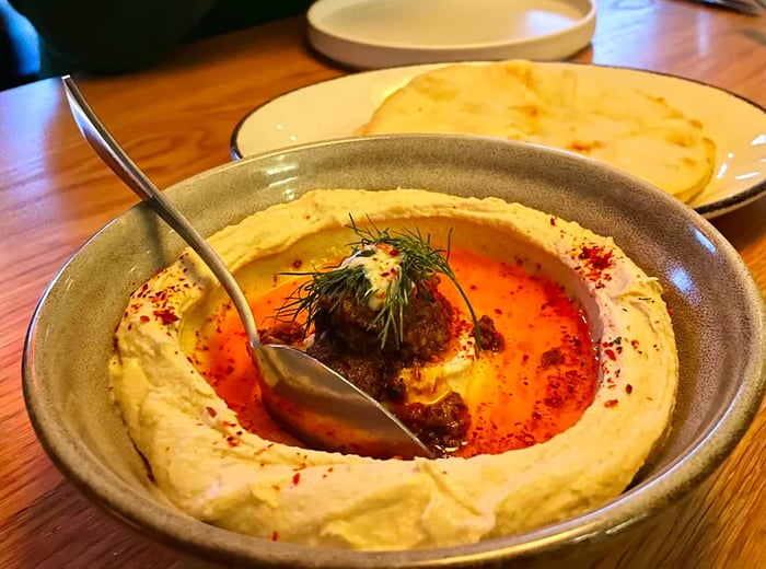 A serving of hummus featuring a central mound of spiced ground meat and hot oil, garnished with herbs and spices, accompanied by a plate of flatbread.