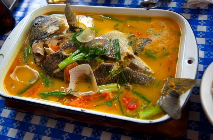 A whole fish, skin intact, is served in a generous baking dish filled with a savory broth, accompanied by hearty chunks of vegetables.