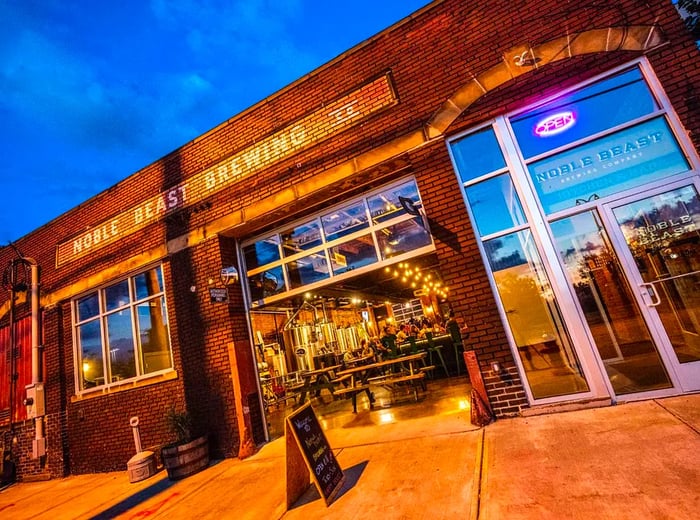 The exterior of a brewpub viewed from a low angle, featuring a brick façade, a large open garage door leading into a spacious interior, and neon signage.