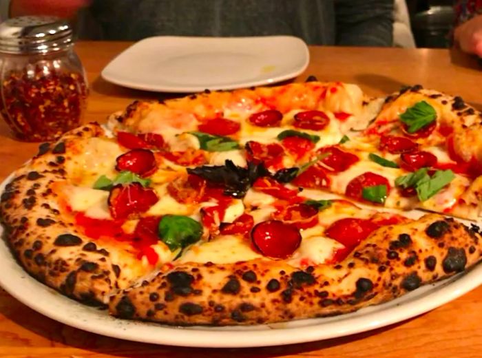 A pepperoni pizza presented on a large platter, topped with fresh basil and a perfectly charred crust, set on a table alongside an empty plate and a shaker of red pepper flakes.