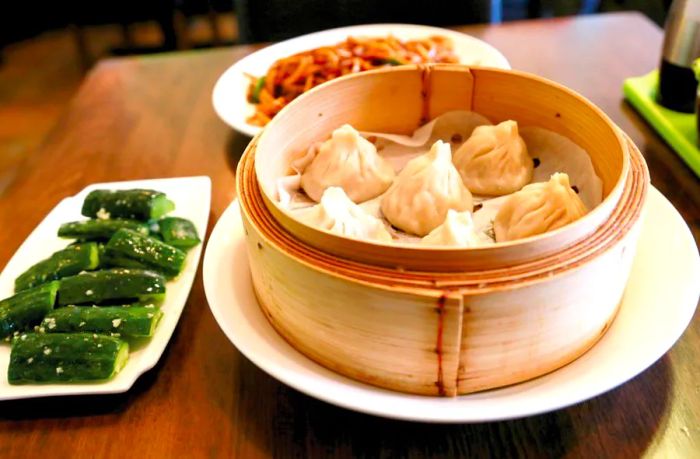 A bamboo steamer filled with soup dumplings next to a plate of cucumbers on a wooden table.