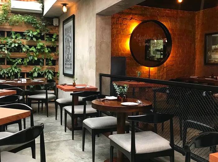 The dining area at Matria features round tables for two with minimalist chairs, a backlit round mirror set in a dark alcove, and natural light pouring in through tinted skylights. The space also boasts a plant wall adorned with cascading vines.