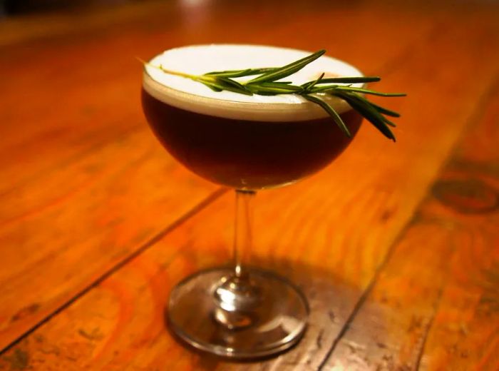 A coupe glass holds a frothy cocktail, elegantly garnished with a sprig of rosemary.