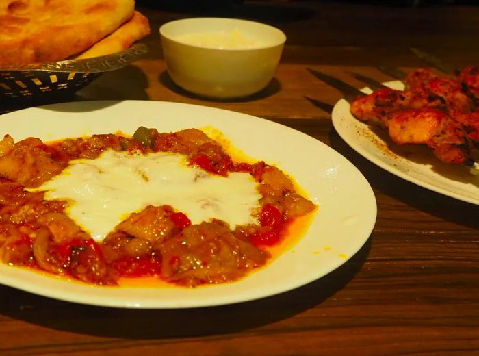 A wooden table is set with a plate of hearty eggplant and tomato stew, skewers, and a small basket of freshly baked flatbreads.