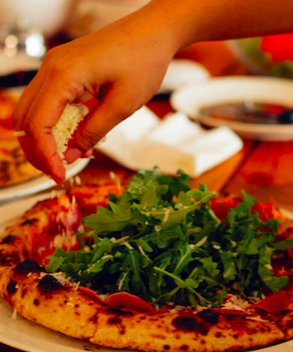 A pizza cook sprinkles grated cheese over a pizza topped with arugula and prosciutto, while other dishes blur into the background on the table.