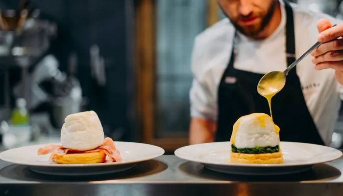 A chef artfully drizzles sauce from a spoon onto a meticulously arranged mound of ingredients on a metal prep table.