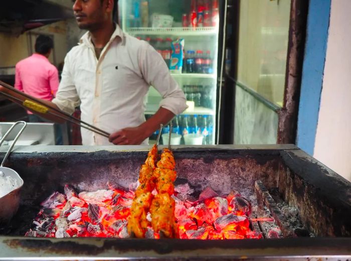 Skewers of marinated chicken sizzle over a charcoal grill, while the vendor readies more skewers in the background.